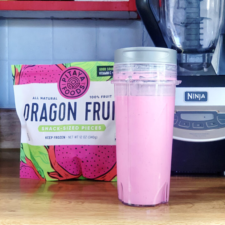 A bright pink dragonfruit smoothie in a plastic tumbler beside a bag of frozen fruit.