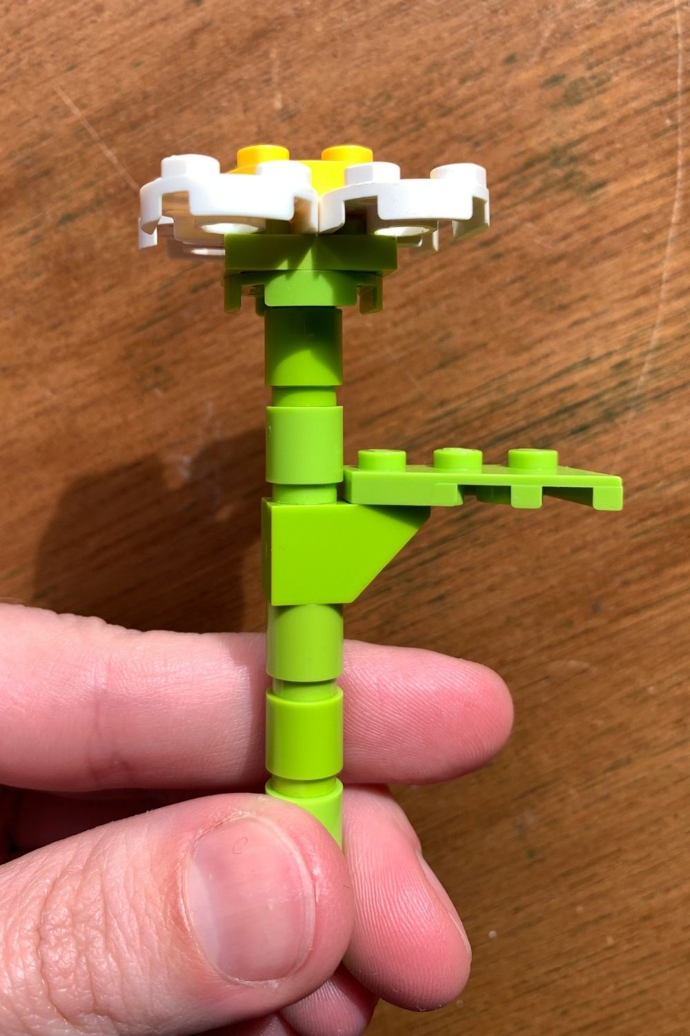 A hand holding a LEGO daisy with a green stem and leaves, white petals, and a yellow center.