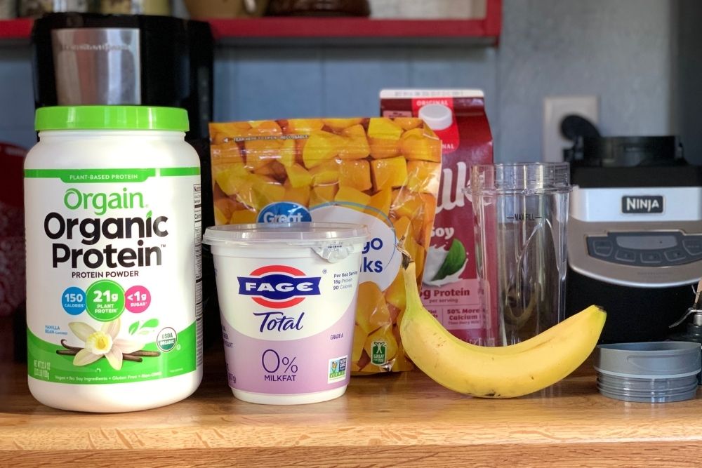 A container of protein powder, yogurt, frozen diced mango, soy milk, and a banana beside a blender.