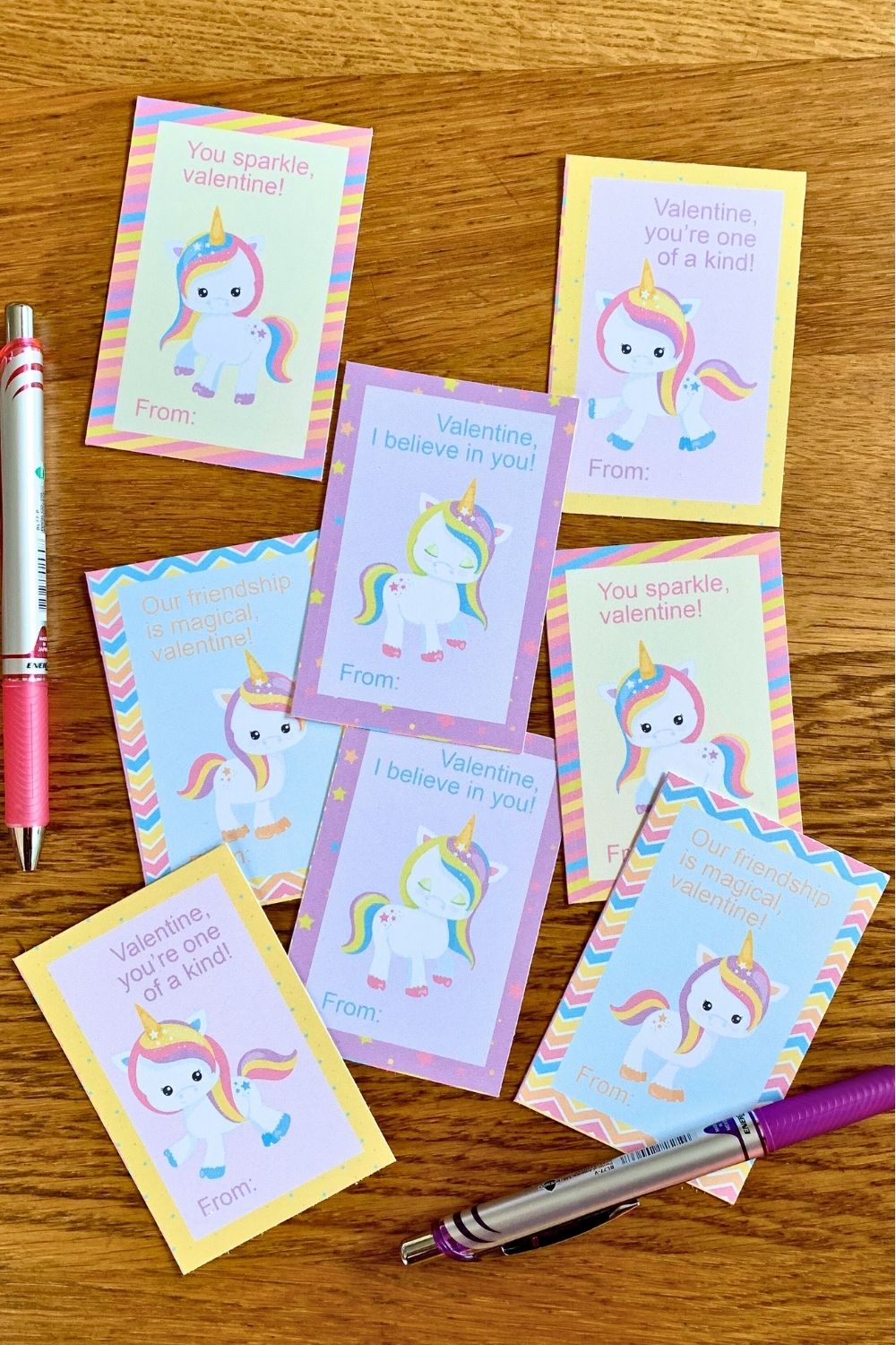 A scattered pile of rainbow unicorn valentine cards beside pink and purple pens.
