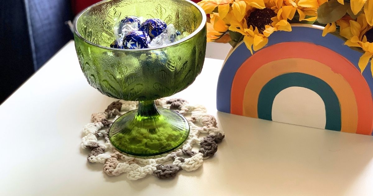 A brown and white crochet doily on an end table under a glass candy dish.