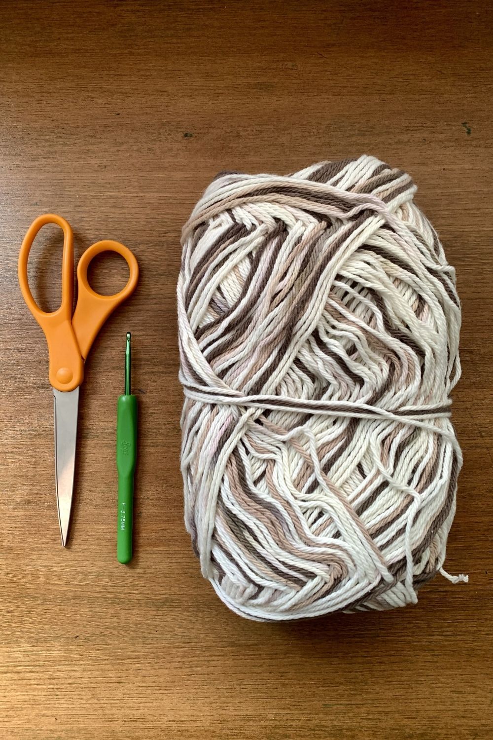 A pair of orange-handle scissors, a size F crochet hook, and a skein of brown and white cotton yarn.