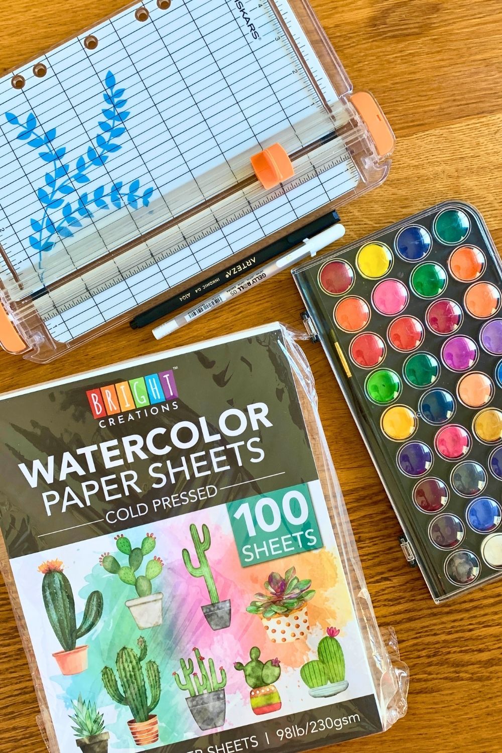 A package of watercolor paper, watercolor paint kit, paper trimmer, and black and white pens.