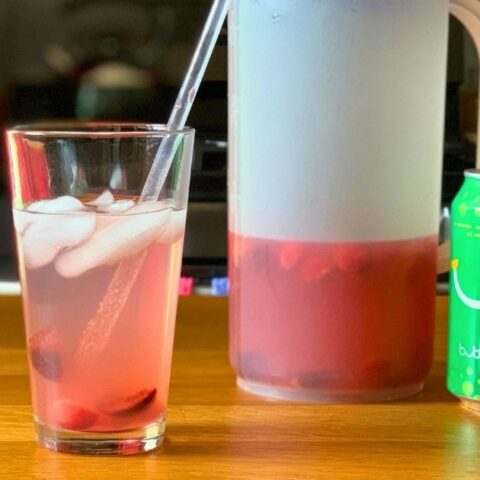 A glass of cherry limeade beside a pitcher of limeade base and a can of sparkling water.