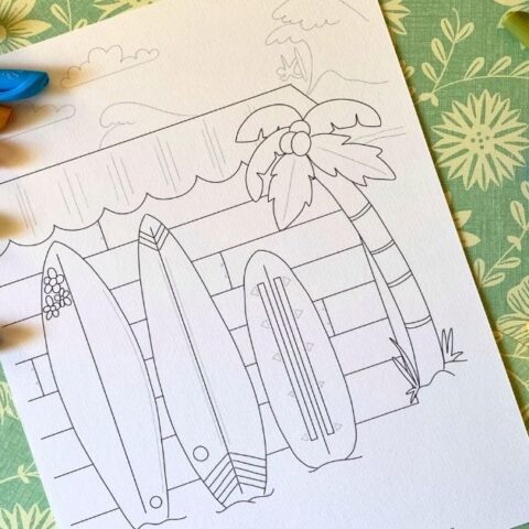 Printable surfboard coloring page with 3 decorated boards leaning against the side of a wood slat building, a surfer catching a wave in the background.