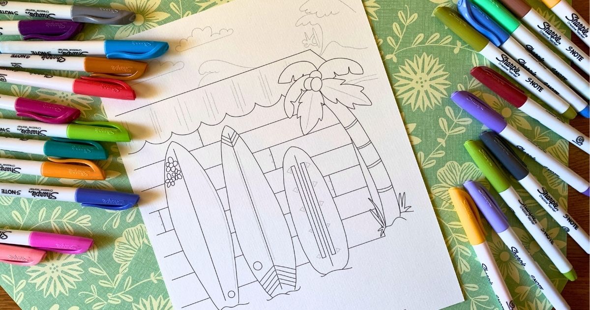 Printable surfboard coloring page with 3 decorated boards leaning against the side of a wood slat building, a surfer catching a wave in the background.