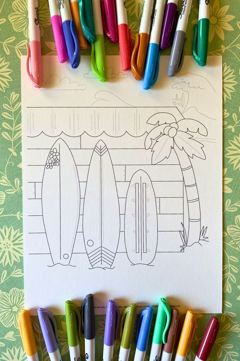 Printable surfboard coloring page with 3 decorated boards leaning against the side of a wood slat building, a surfer catching a wave in the background.