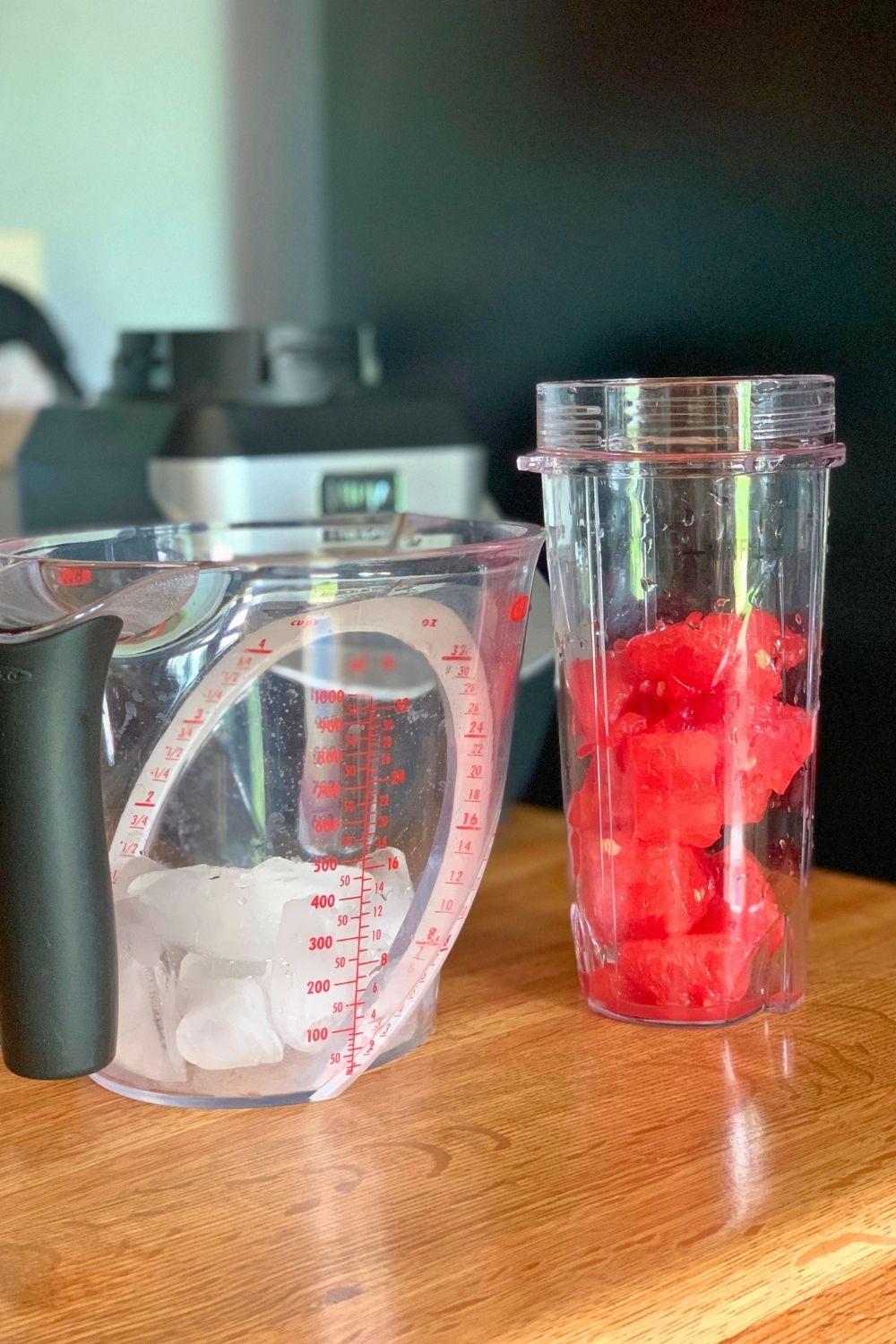 A blender cup of watermelon chunks beside a measuring cup with ice cubes.