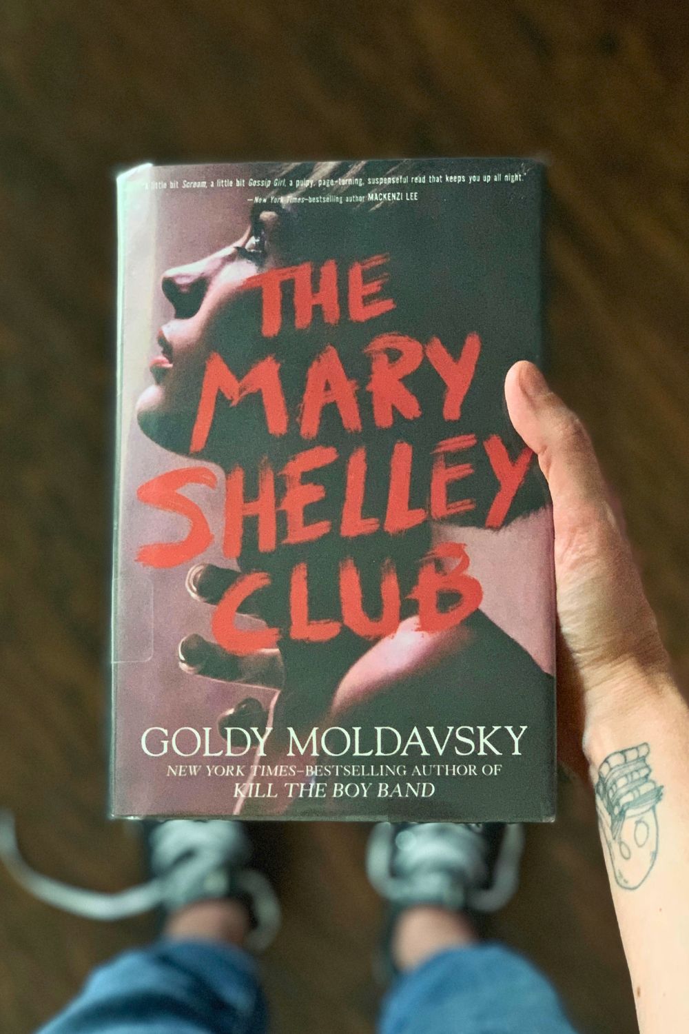 A hand holding the book The Mary Shelley Club, by Goldy Moldavsky, above feet wearing untied black Converse sneakers.