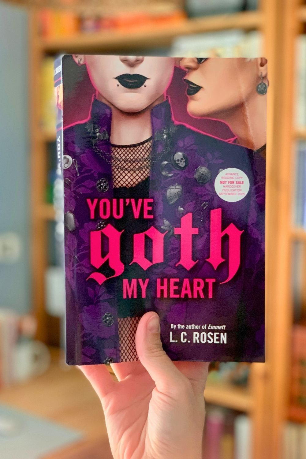 A hand holding the book You've Goth My Heart, by Lev AC Rosen, in front of a full bookshelf.