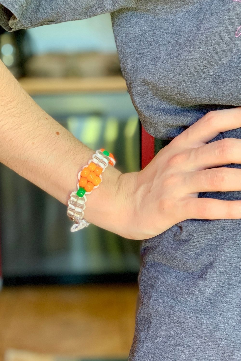 A person wearing a pumpkin bracelet made from orange, green, and clear pony beads on cord.