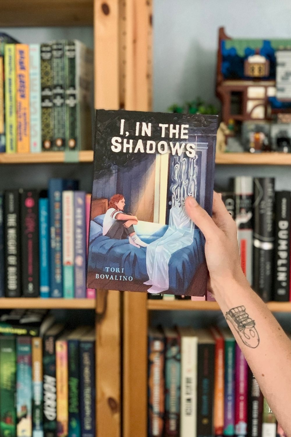 A hand holding a hardcover book, I In the Shadows by Tori Bovalino, in front of a bookshelf.