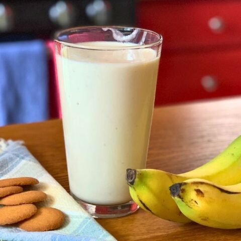Banana cream pie smoothie in a glass beside a whole banana and a stack of vanilla wafers.