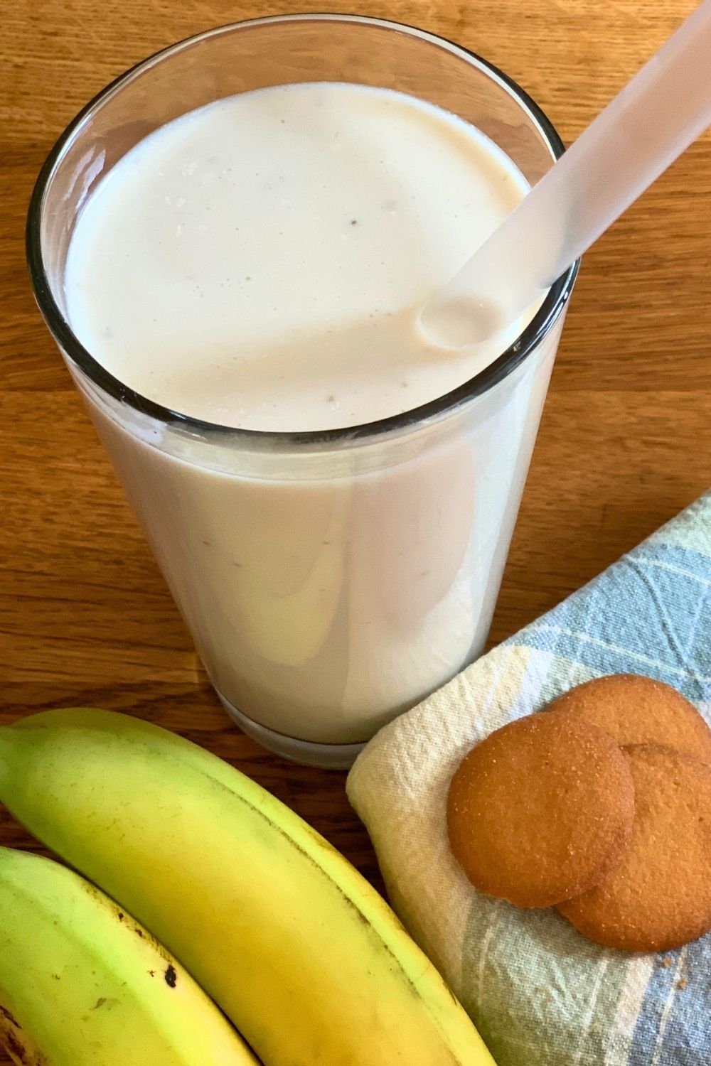 Banana cream pie smoothie in a glass beside a whole banana and a stack of vanilla wafers.