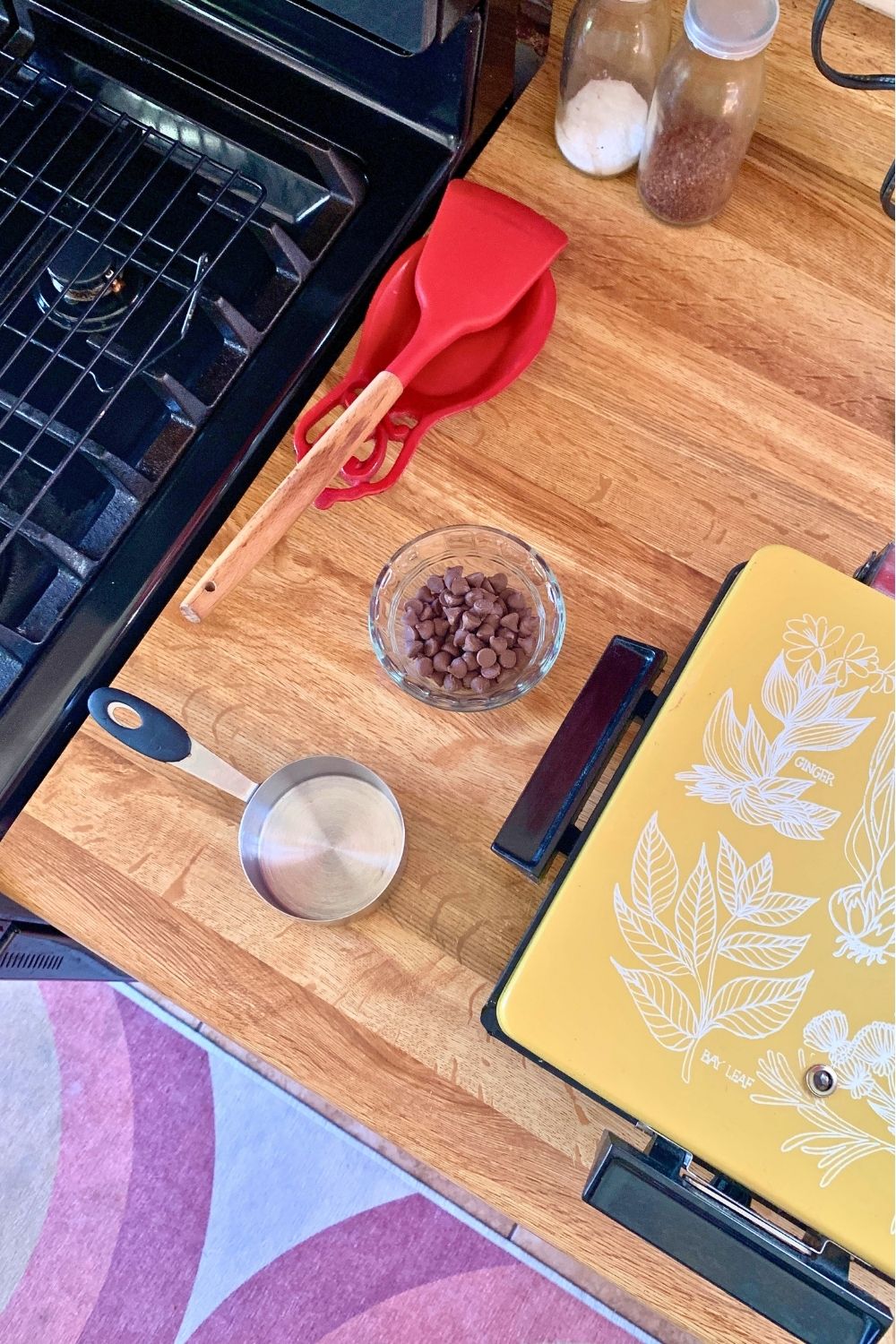 A closed waffle iron beside a measuring cup, bowl of chocolate chips, and spatula.
