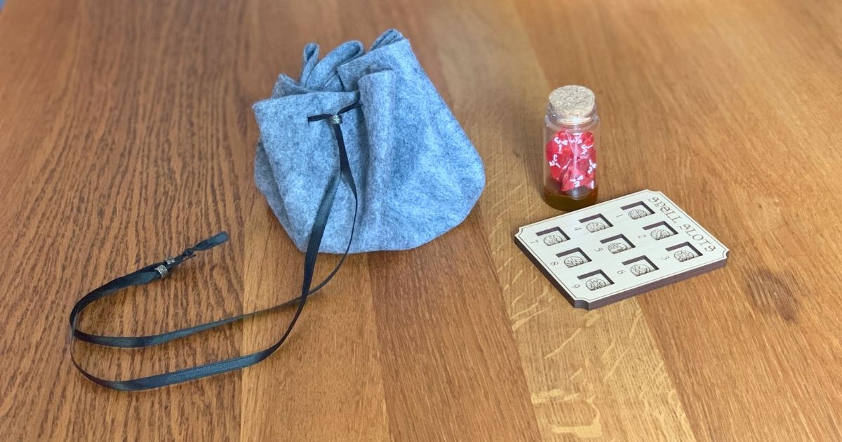 A gray felt dice bag with a black ribbon drawstring beside a jar of red dice and a spell slot card.