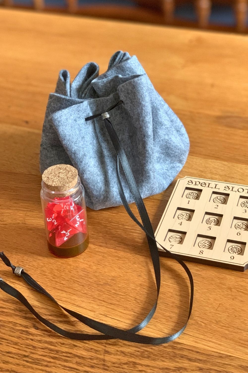 A gray felt dice bag with a black ribbon drawstring beside a jar of red dice and a spell slot card.