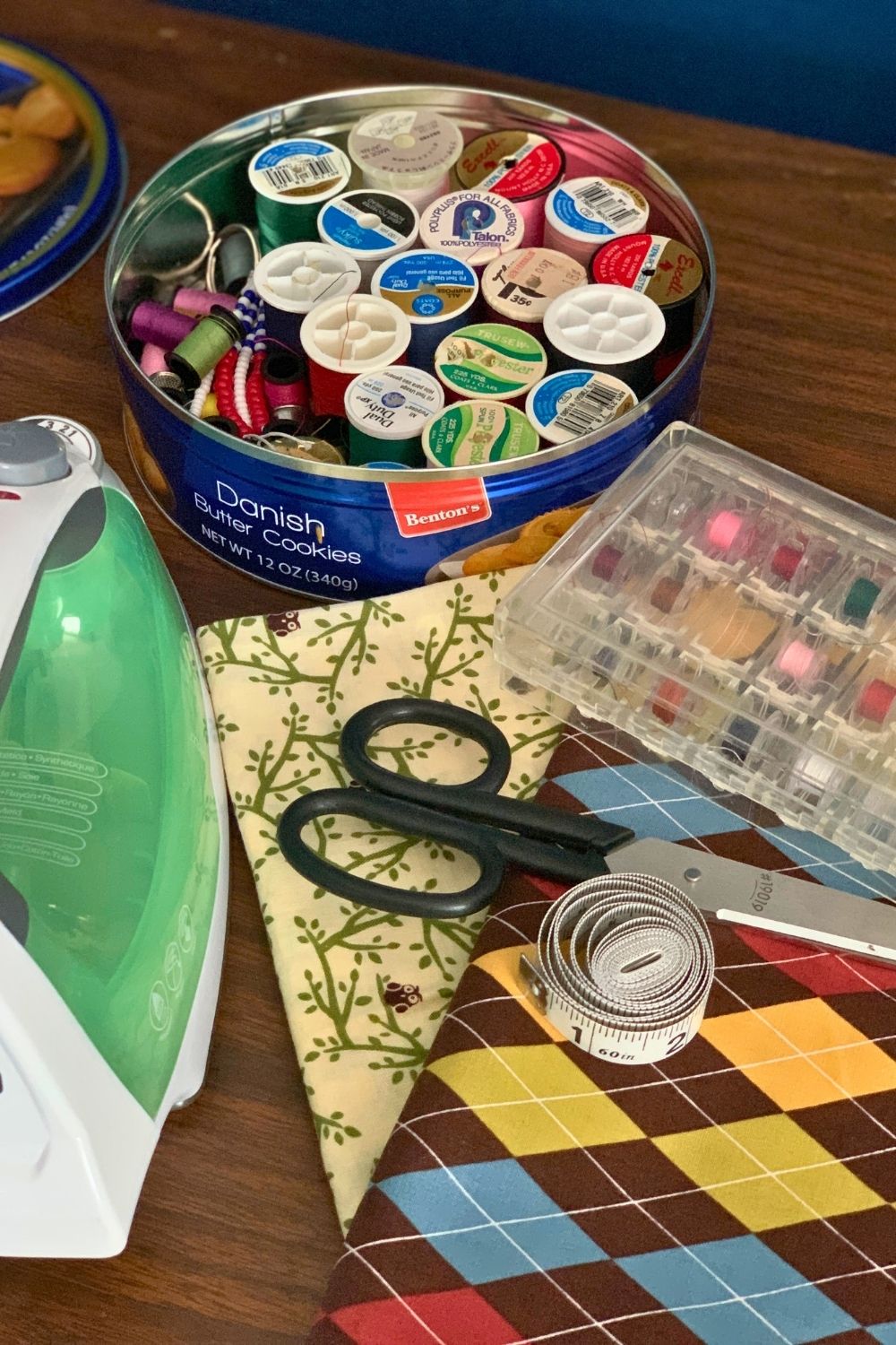 Two patterned fabrics, scissors, tape measure, an iron, bobbins, and a tin of thread.