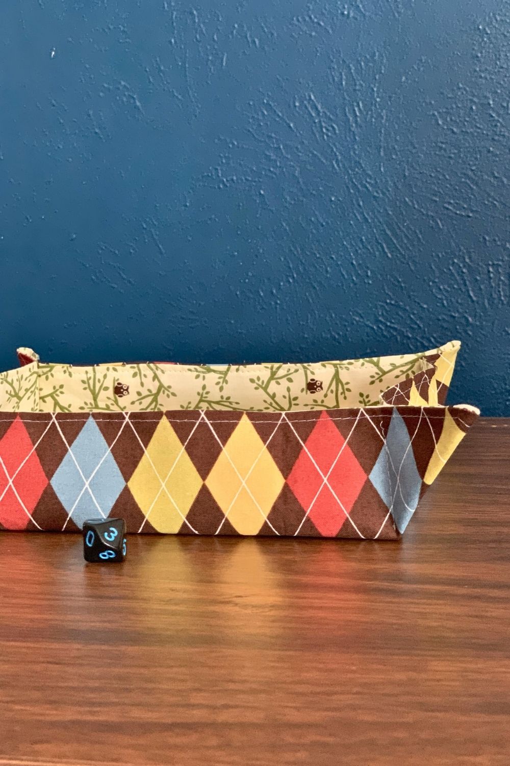 A square fabric tray with red, blue, yellow, and brown argyle pattern outside and owns and leaves inside.