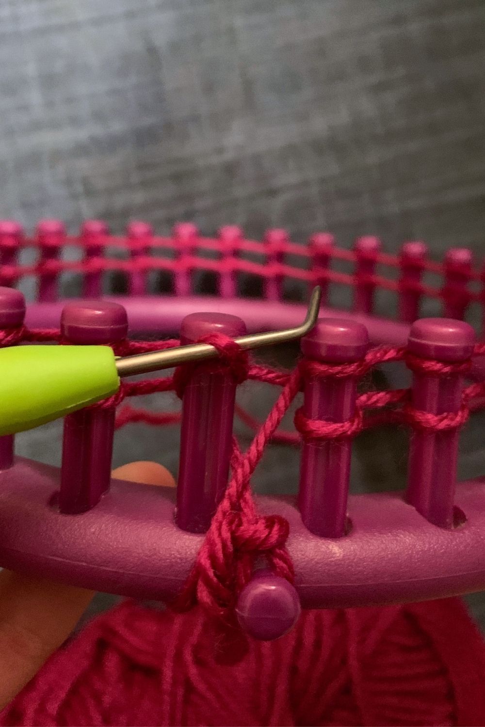 Using a knitting loom hook to lift a lower loop over an upper loop and peg.