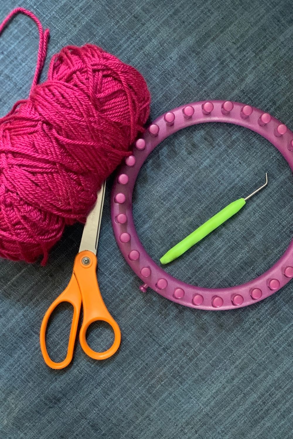 A skein of fuchsia yarn, pair of scissors, knitting loom, and hook.