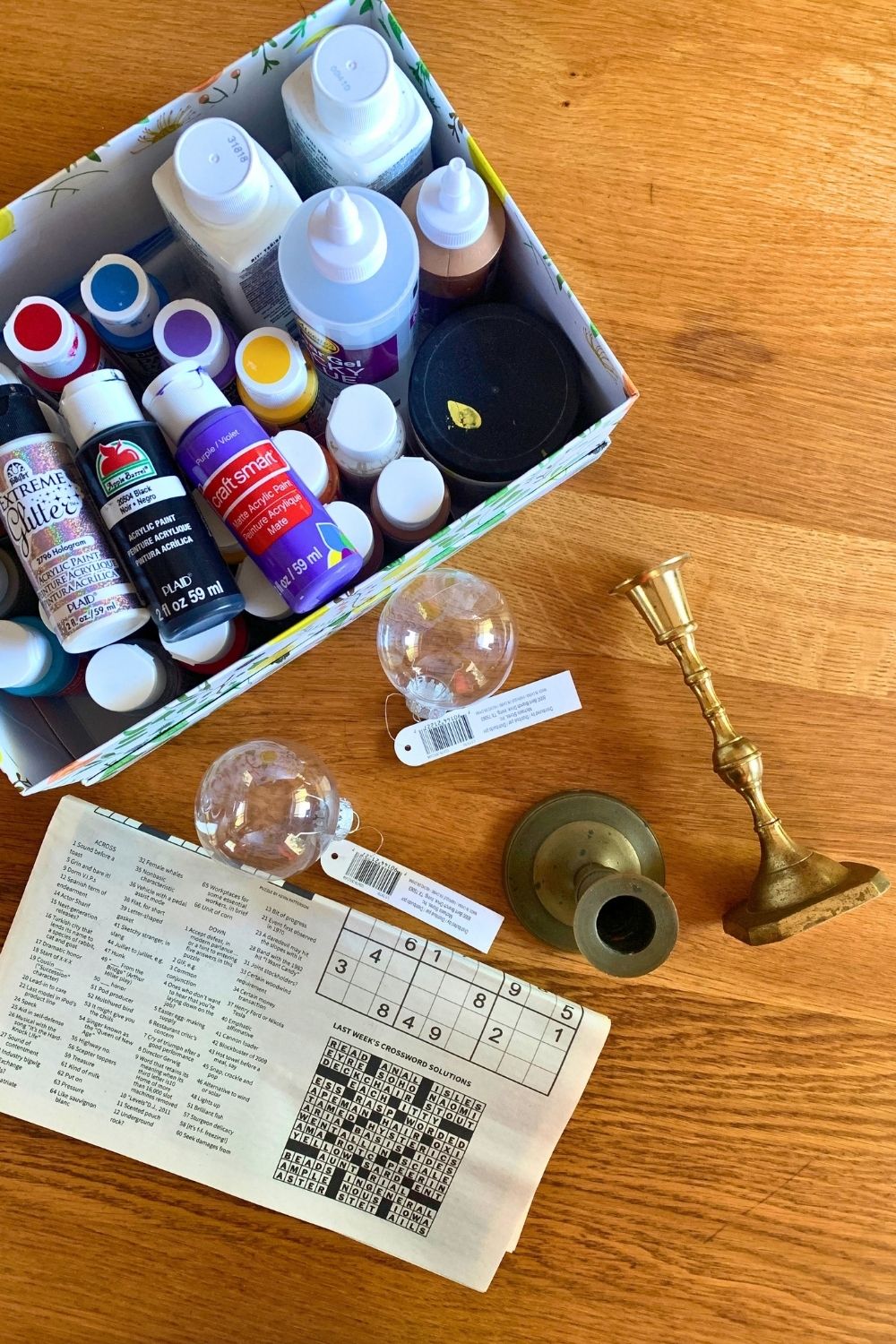 A shoebox of several small paint bottles, a folded newspaper, brass candlestick holders, and 2 clear ball ornaments.