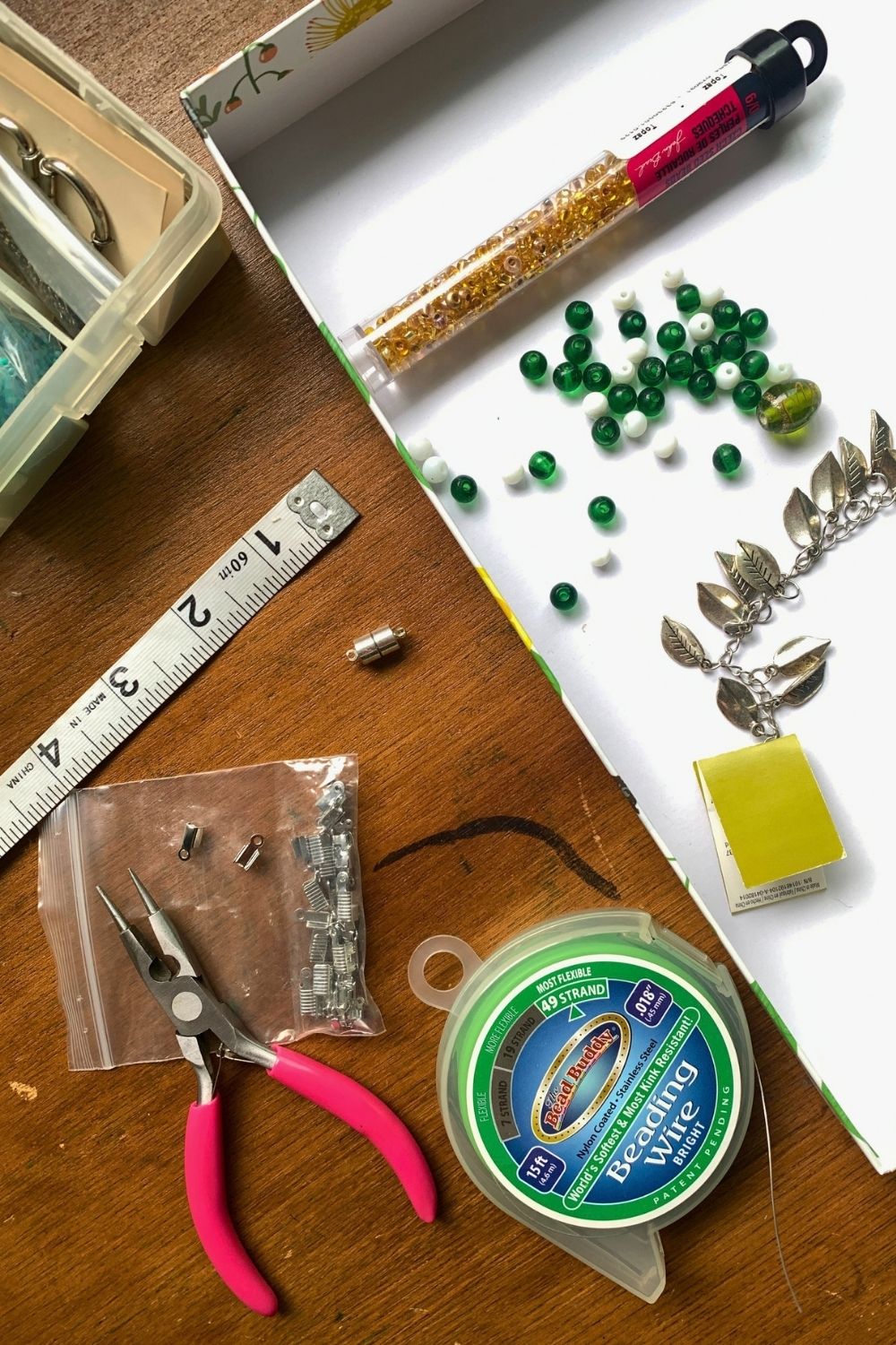 A soft tape measure, jewelry pliers, glass beads in green, white, and gold, silver leaf charms, roll of flexible beading wire, silver end crimps, and a silver magnetic closure spread on a work table.