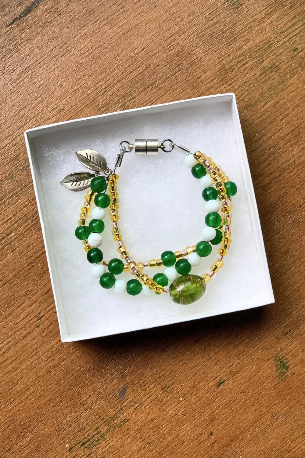 A handmade bead bracelet with 3 strands of glass beads in green, white, and gold patterns and a pair of leaf charms at the clasp.