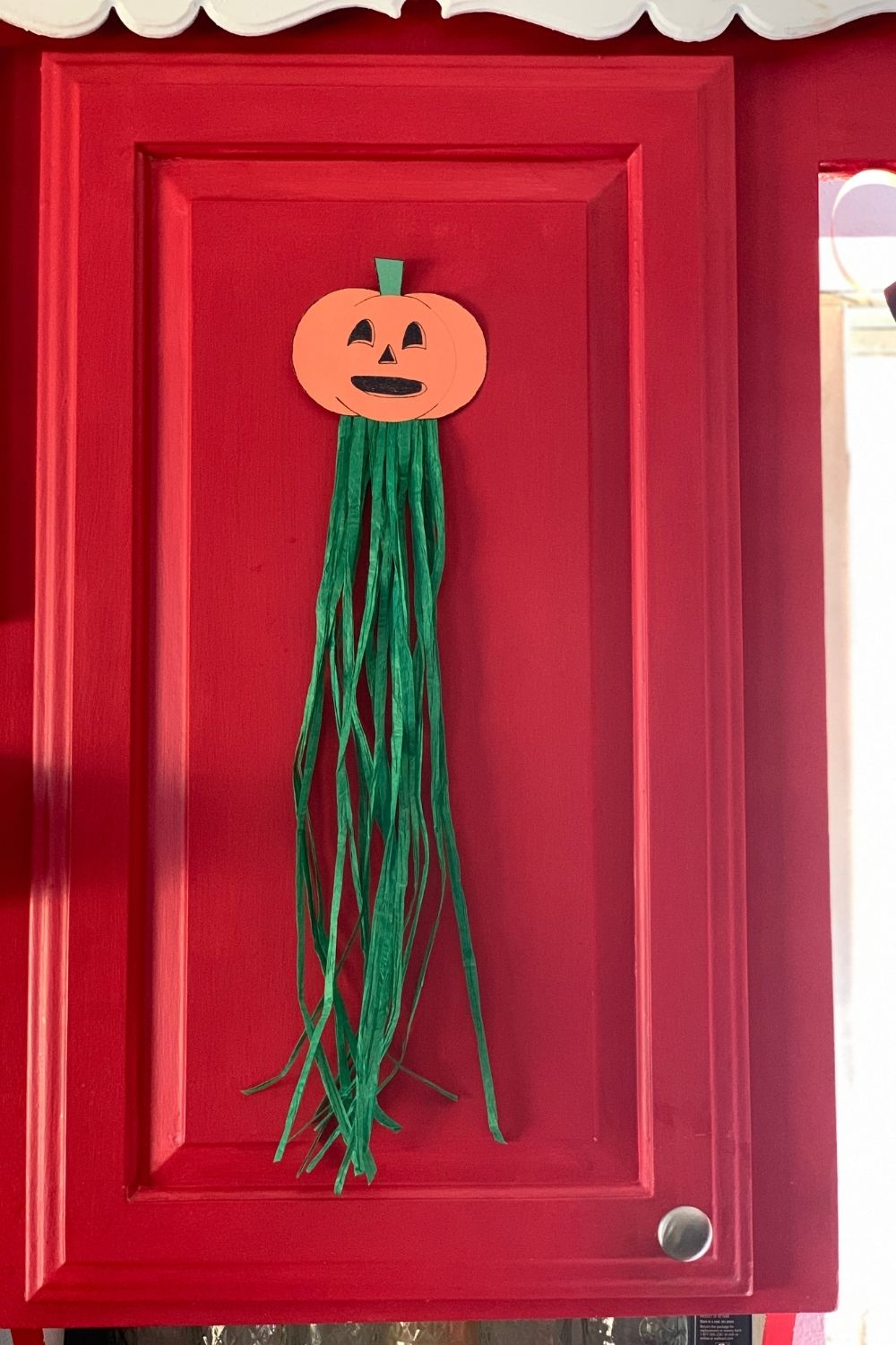 An orange paper jack-o'lantern with green raffia streamers stuck to a kitchen cabinet door.