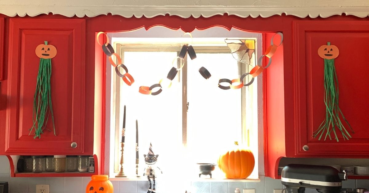 A kitchen decorated for Halloween with a pair of paper pumpkins with green raffia tassels stuck to cabinet doors, paper chains, with a retro plastic jack o-lantern, cat, and candles.