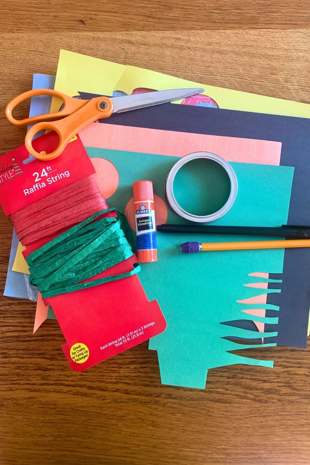 A stack of multicolor construction paper, scissors, glue stick, green raffia roll, pencil, black pen, and canning jar ring.