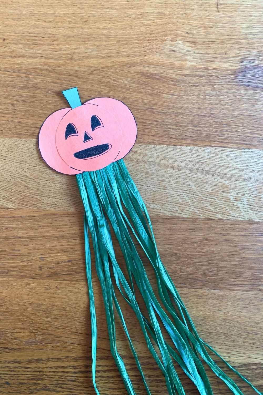 An orange construction paper jack-o'lantern with a green stem and long green raffia tassels.