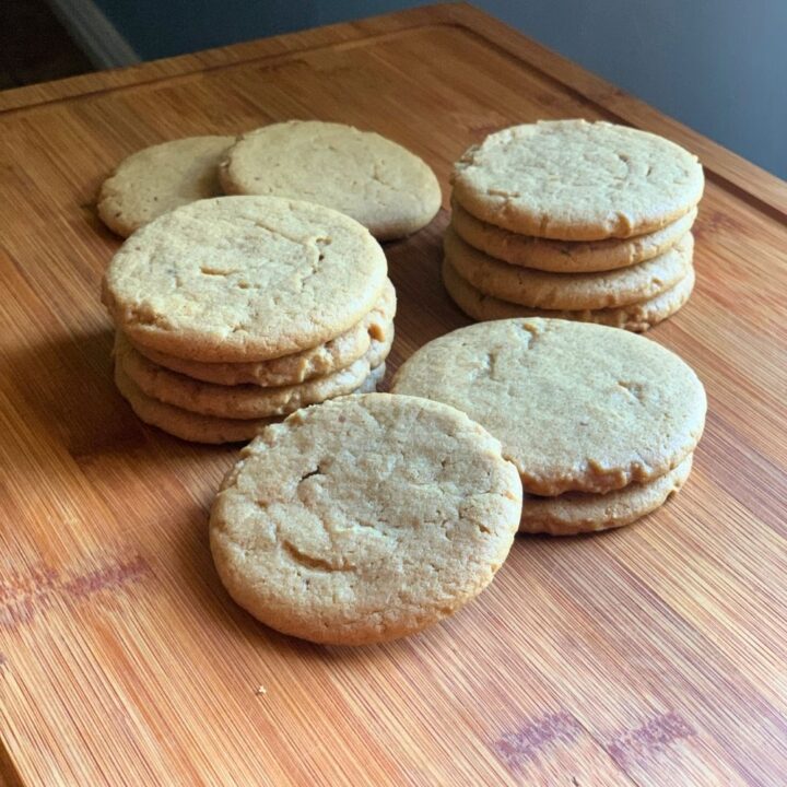 Several stacks of light brown peanut butter cookies on a wood cutting board.