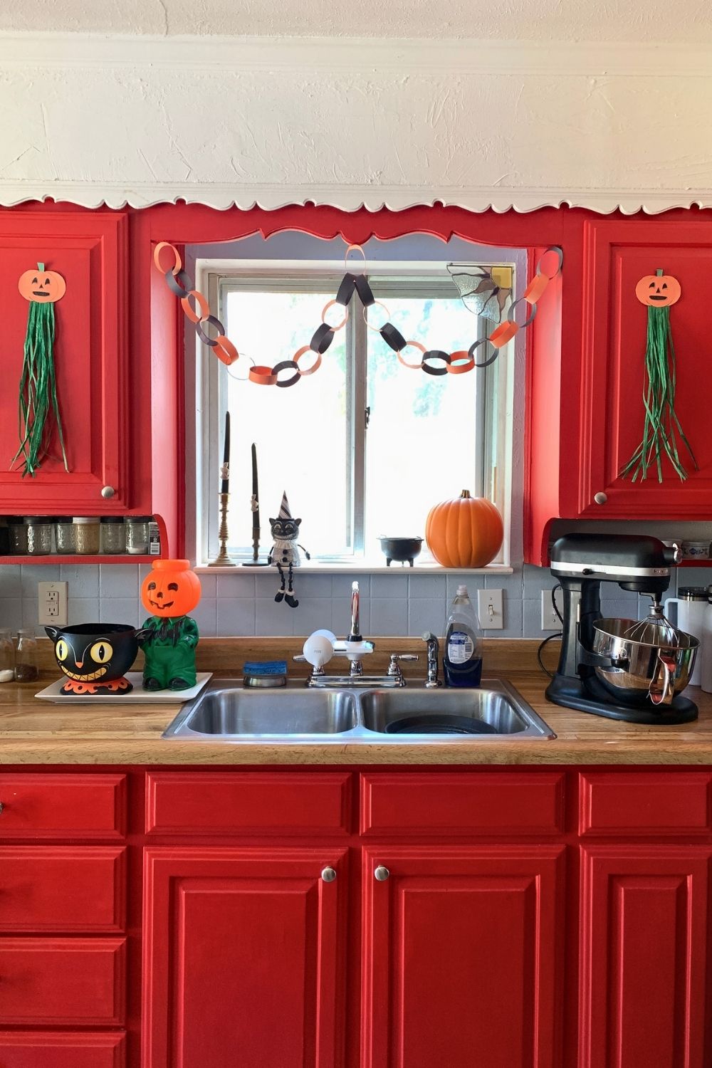A kitchen decorated for Halloween with a pair of paper pumpkins with green raffia tassels stuck to cabinet doors, paper chains, with a retro plastic jack o-lantern, cat, and candles.