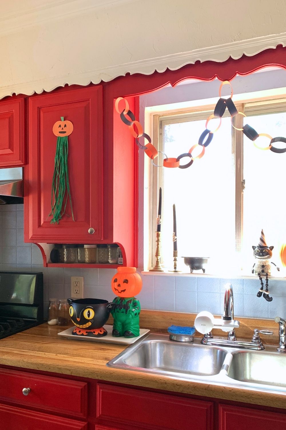 A kitchen decorated for Halloween with a pair of paper pumpkins with green raffia tassels stuck to cabinet doors, paper chains, with a retro plastic jack o-lantern, cat, and candles.